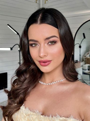 Woman with wavy brown hair, soft glam makeup, and neutral tones.