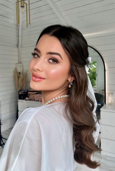 Woman with wavy brown hair, soft glam makeup, and neutral tones.